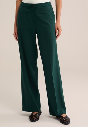 Femme portant un pantalon vert foncé à jambes larges et des chaussures noires tissées sans lacets, debout sur un fond clair.