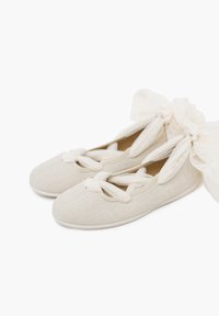 Light beige ballet flats feature a textured upper and decorative bow on each side, resting on a white sole against a plain background.