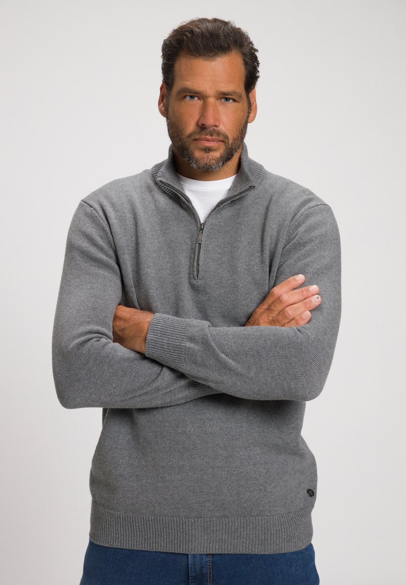 Mann mit dunklen Haaren und Bart, der einen grauen Halbzip-Pullover und blaue Jeans trägt, steht mit verschränkten Armen vor einem schlichten Hintergrund.