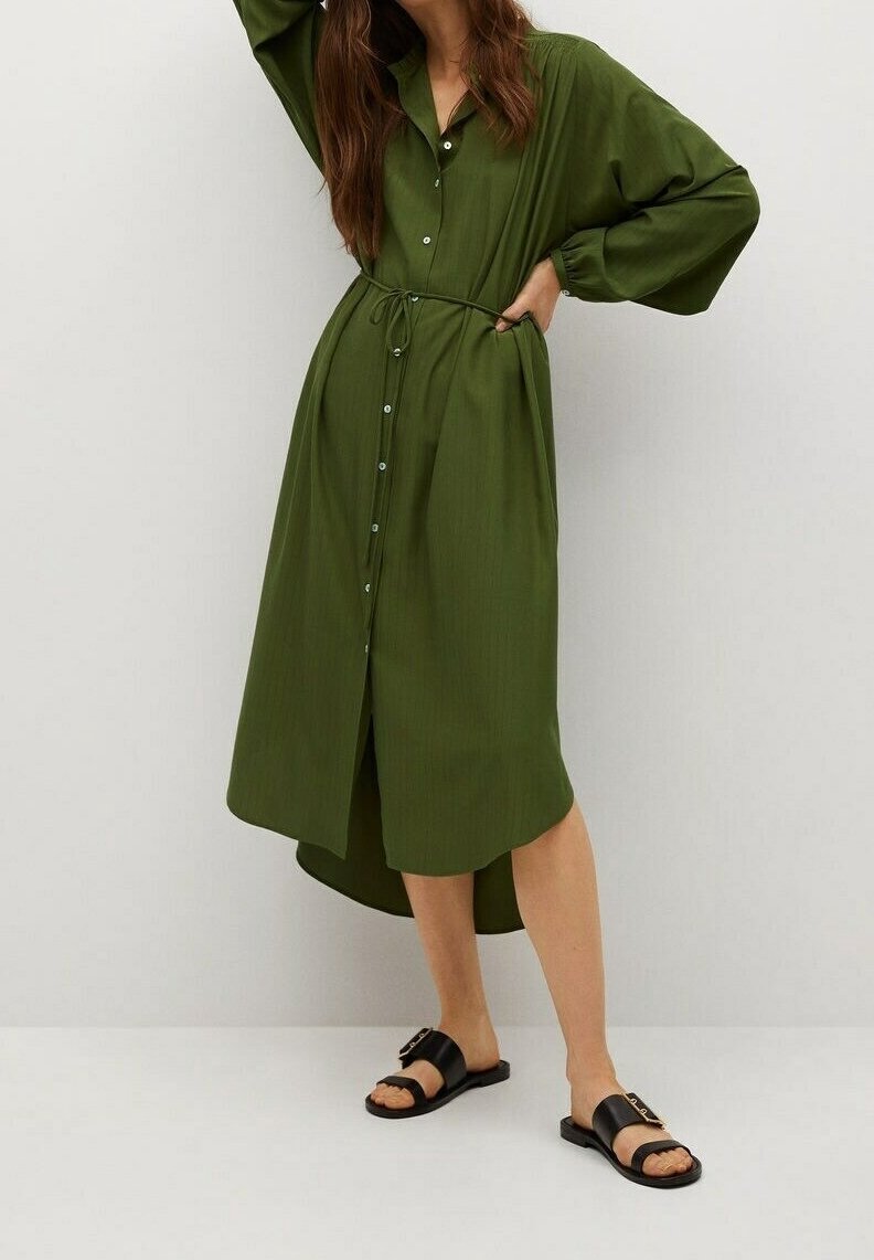 Femme portant une robe boutonnée longue manche vert olive avec une taille nouée et des sandales plates noires sur un fond clair uni.