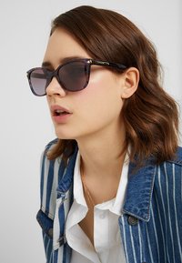 Ochelari de soare cu carapace de broască țestoasă, cu lentile închise; brațele negre au logo-ul auriu "COACH". Modelul poartă o jachetă din denim cu o cămașă albă dedesubt.