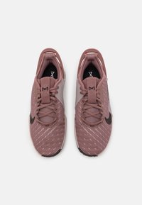 Zapatillas deportivas de color rosa claro, hechas de malla y materiales sintéticos. Presentan un logo negro, una parte superior texturizada y un sistema de cierre con cordones y talón reforzado.