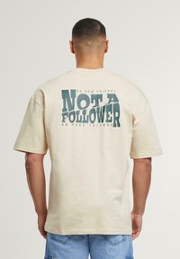 Beige cotton T-shirt with oversized fit. Graphic on the back features bold green text: "NOT A FOLLOWER," with smaller text above and below.