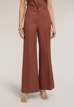 Mujer con pantalones de tiro alto y pernera ancha de color óxido con bolsillos frontales y sandalias doradas de tacón alto con tiras, de pie frente a un fondo liso.