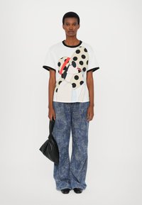 White t-shirt with a colorful graphic of a woman wearing a polka dot hat, paired with blue patterned wide-leg jeans and a black handbag.