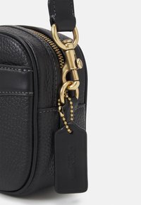 Borsa in pelle nera texturizzata con cerniera e dettagli in oro. Presenta un attacco a catena con un'etichetta nera che mostra il nome del marchio.