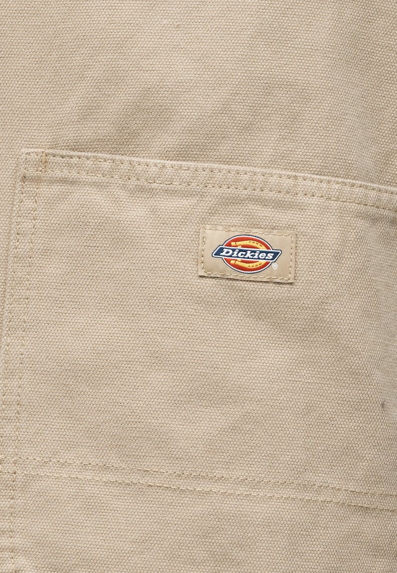 Tela de lona beige con un bolsillo cuadrado que incluye un pequeño parche rectangular con el logotipo de la marca Dickies cosido.