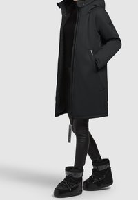 Personne portant un long manteau d'hiver noir avec des poches zippées, un pantalon noir et des bottes de neige matelassées noires avec le logo "Moon Boot".