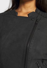 Giacca in ecopelle nera con colletto alto, cuciture dettagliate e diverse tasche con zip per lo stoccaggio. Texture liscia e design moderno.