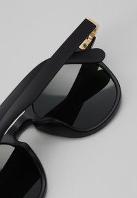 Black sunglasses with a matte finish, wide rounded lenses, gold hardware accents at the hinge, and curved temple arms.