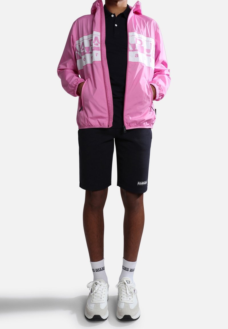 Coupe-vent rose avec des accents blancs, fermeture éclair, poches latérales, porté par-dessus un polo noir, associé à un short noir et des baskets blanches.