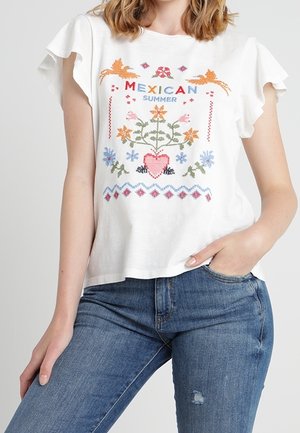 Blouse blanche à manches courtes avec manches volantées, broderies florales et cœurs colorés, avec le texte "Mexican Summer", associée à un jean bleu.