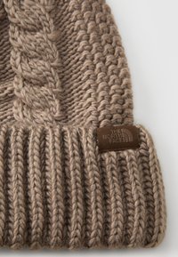 Gorro de malha castanho com textura canelada e design de trançado, apresentando uma etiqueta de tecido castanha com o logotipo "The North Face".