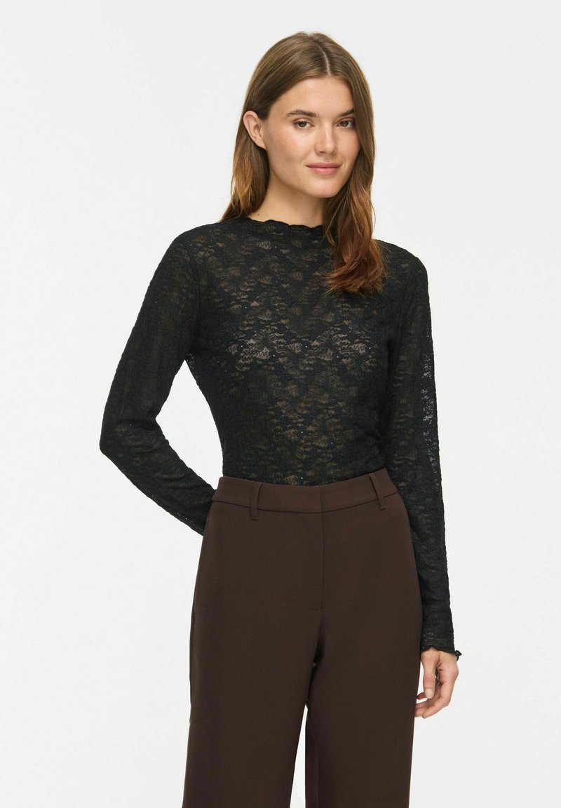 Femme aux cheveux châtain clair portant un top en dentelle noir à manches longues et un pantalon taille haute marron foncé, debout avec une main dans le dos.