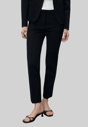 Black tailored trousers featuring a slim fit, smooth texture, and front pleats, paired with black sandals with a decorative strap.