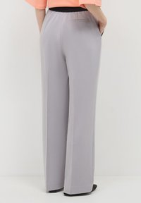 Personne portant un pantalon large gris clair et un haut pêche, se tenant avec les mains dans les poches devant un fond uni.