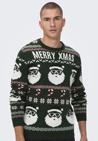 Strikket julesweater, grønn med hvite nisseansikter, røde julegodterstokker og snøfnugg. Teksten lyder "GOD JUL." Ribbet rund hals og mansjetter.