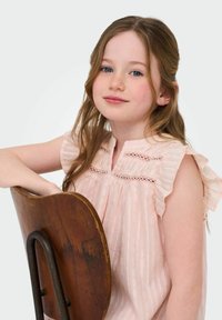 Ljusrosa blus med volangärmar och spetsdetaljer, bärs av en flicka som vilar sin hand på en trästol med en böjd rygg.