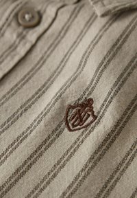 Stripete beige skjorte i stoff med en brun brodert logo og knappedetaljer, som fremhever et strukturert, vevd utseende.