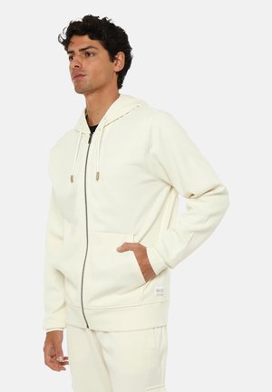 Uomo che indossa una felpa crema con zip e coulisse e pantaloni coordinati, in piedi con una mano in tasca su uno sfondo semplice.