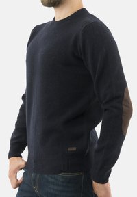 Mężczyzna w granatowym swetrze z brązowymi łaty na łokciach i ciemnych dżinsach, stojący na białym tle.