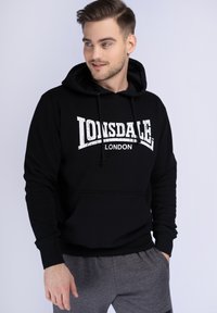 Čierna mikina s kapucňou a bielym potlačou "Lonsdale London". Obsahuje klokanie vrecko a rebrované manžety. Nosená s šedými teplákovými nohavicami.