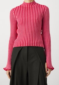 Czerwono-różowy sweter w paski z ribbkontowym splotem, z wysokim kołnierzem i rozkloszowanymi mankietami, wykonany z teksturowanego materiału. Połączony z ciemnymi spodniami.