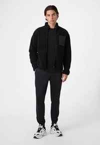 Schwarze Fleecejacke mit Reißverschluss, ausgestattet mit einer Brusttasche und kombiniert mit schwarzen Jogginghosen und weißen Sneakers, die kontrastreiche Akzente setzen.