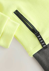 Neon żółty skrócony sweatshirt z czarną kieszenią na zamek i metką BAKER. Rękaw ma miękką fakturę z kontrastującym ciemnoszarym mankietem.