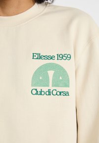 Krémová mikina s kapucí se zeleným vyšitým textem "Ellesse 1959" a kulatou grafikou s linkami nad "Club di Corsa."