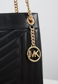 Sac à main en cuir noir avec motif matelassé, bandoulière en chaîne dorée et pendentif rond en or avec le logo Michael Kors suspendu à la poignée.