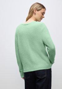 Pull en menthe verte, texturé, avec une coupe décontractée, col rond et manches longues, associé à un pantalon en denim foncé. Fond clair.