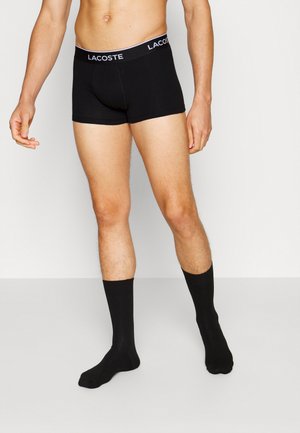 Black boxer shorts with a contrasting white “LACOSTE” logo band at the waist, paired with black ankle-length socks. Smooth fabric, fitted design.