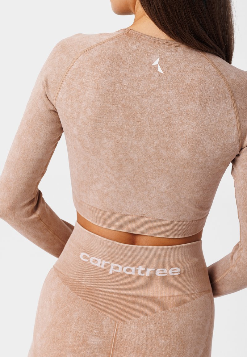 Jasnobrązowa sportowa bluzka z długim rękawem, z prążkowanymi mankietami i logo z tyłu. Połączona z dopasowanymi legginsami z wysokim stanem, które mają białe napisy marki.