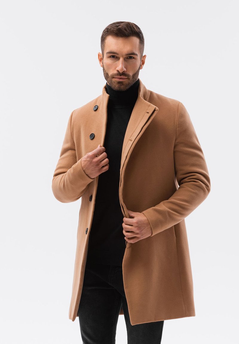 Cappotto di lana beige con un design aderente, dotato di un colletto alto, bottoni neri e una texture liscia, indossato sopra un dolcevita nero.