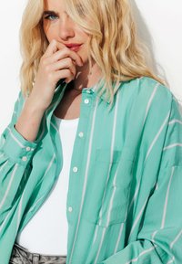 Chemise boutonnée turquoise avec des rayures verticales roses, coupe décontractée, poche poitrine et manches longues. Portée par-dessus un haut blanc.