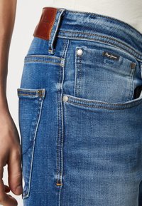 Acerca de los jeans de mezclilla azul en primer plano, mostrando los bolsillos delanteros y laterales, remaches metálicos, costuras en contraste y una etiqueta de marca de cuero marrón en la cintura.