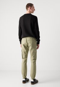 Zwarte trui, licht olijfgroene cargobroek met elastische boorden en zijzakken. Schoenen zijn zwart met witte en bruine accenten.
