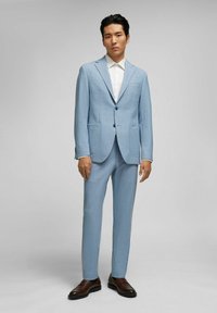 Lichtblauw pak met een gestructureerde stof, inclusief twee voorzakken en een enkele knoopsluiting, gecombineerd met een witte overhemddraag en bruine schoenen.