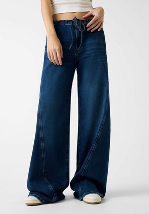 Jean en denim à jambe large de couleur bleu foncé, avec une taille à cordon, des coutures latérales et une texture lisse. Associé à des chaussures blanches.