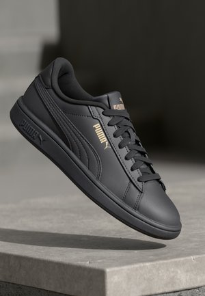 Zwarte Puma sneaker met leren bovenkant, gouden logo en zwarte veters, zwevend boven een stenen oppervlak tegen een donkere achtergrond.