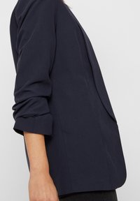 Námořnicky modré sako s upraveným střihem, hladkou tkaninou, ohrnutými manžetami a postranními rozparky a kapsami, které představují moderní siluetu.