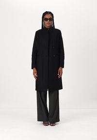 Cappotto nero doppiopetto realizzato in un tessuto morbido, con maniche lunghe e una silhouette dritta, abbinato a pantaloni a gamba larga con righe verticali.