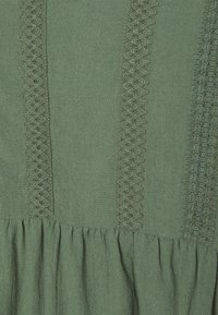 Vihreä kangas, jossa on teksturoituja raitoja ja kerätty helma. Sisältää koristeompelua pystysaumojen varrella, mikä luo kerroksellisen ulkonäön.