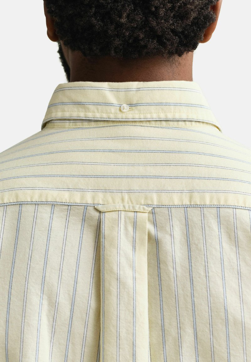 Vista trasera de una persona que lleva una camisa amarillo pálido con finas rayas verticales y horizontales azules, mostrando los detalles del cuello y del canesú de la camisa.