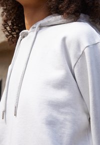 Biała bluza z kapturem uszyta z miękkiego materiału, wyposażona w sznurek ze metalowymi końcówkami oraz luźny krój. Tekstura wydaje się gładka i przyjemna w dotyku.