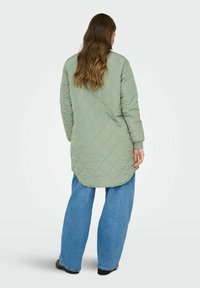 Gaiši zaļa iešūta jaka ar augstu un zemu apdari un garām piedurknēm, apvienota ar plašu zilu džinsu biksēm. Gluda tekstūra, minimālistisks dizains.