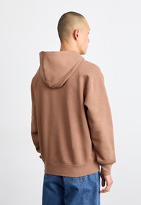 Sweat-shirt à capuche marron en tissu doux avec des poignets et un ourlet côtelés, associé à un jean bleu. La vue de dos montre la capuche et la coupe décontractée.