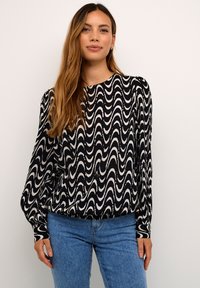 Schwarz-weiße Bluse mit langen Ärmeln, die ein wellenförmiges Muster, einen runden Ausschnitt und eine lockere Passform aufweist, kombiniert mit hellblauen Denim-Jeans.