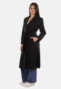 Donna in piedi che indossa un lungo cappotto nero con cintura, jeans larghi blu e sneakers bianche, posa con una mano nella tasca del cappotto.